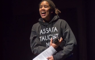 The first ever PURPOSE team member (Nia) stands in front of a black background with eyes squinting, mouth open in laughter. Yeye wears a black hoodie that reads "ASSATA TAUGHT ME" in thin white print. Their arms curl as if holding a baby with a phone in one hand and a paper program in the other. Photo by Charles R. Berenguer, Jr. - Ibaye