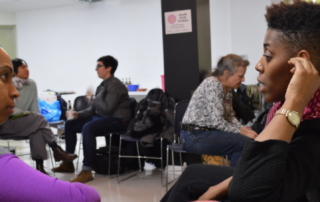 T.L. Daniels, wearing a black long-sleeved top and a patterned red scarf, sits in a dialogue with Sarita, a workshop participant wearing a short-sleeved gray top with longer purple sleeves under it. In the background are other seated participants.